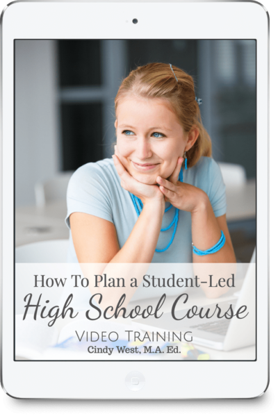 Blonde girl with blue eyes, wearing a blue shirt and blue jewelry smiles and stares into the distance with her elbows on the table beside her computer.